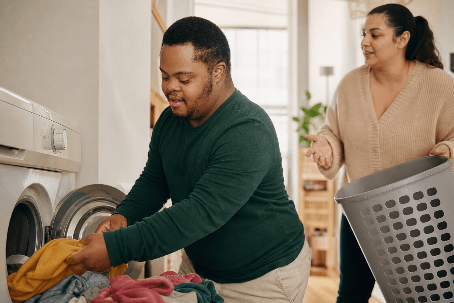A Black man with Down syndrome loads his washing machine while a support professional coaches nearby.